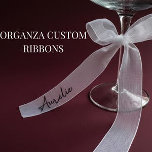 Puede incluir: Una cinta de organza blanca atada alrededor de una copa transparente con un lazo. La cinta tiene la palabra "Aurélie" impresa en escritura negra. El texto "ORGANZA CUSTOM RIBBONS" está en mayúsculas blancas.