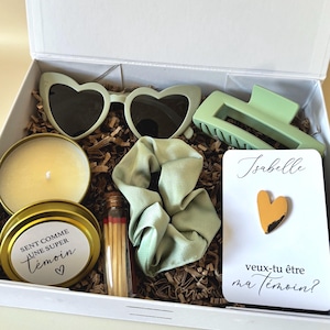 Può includere: Una confezione regalo bianca con vari oggetti: occhiali da sole a forma di cuore, una candela, un elastico per capelli, una clip per capelli, fiammiferi e un biglietto. Il biglietto dice "Isabelle, veux-tu être ma témoin?" e presenta una spilla a forma di cuore. Gli oggetti sono in tonalità verdi e oro.