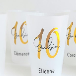 Può includere: Bicchieri di plastica trasparente con numeri e nomi in foglia d'oro. I bicchieri riportano "10" e "Gabin" con i nomi "Clemence", "Etienne" e "Coralie" sotto.