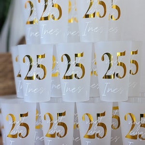 May include: A stack of frosted plastic cups with gold lettering. The cups are decorated with the number "25" and the word "vingt-cinq" in French. The name "Ines" is written on each cup.