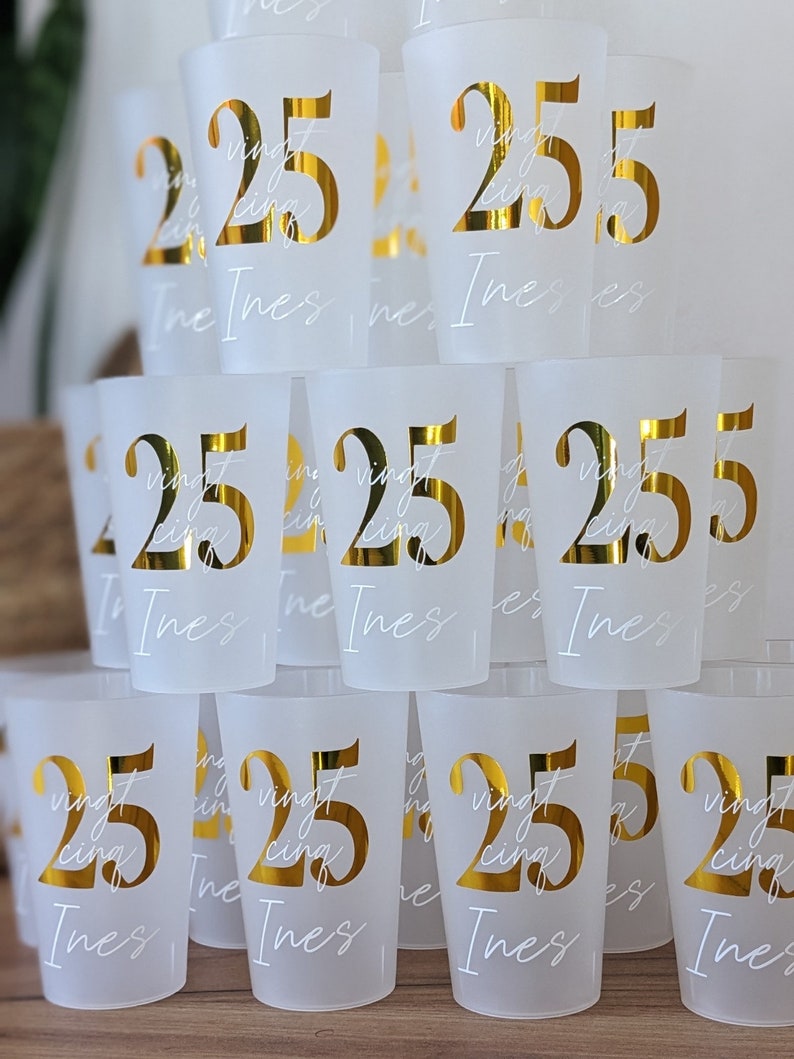 May include: A stack of frosted plastic cups with gold lettering. The cups are decorated with the number "25" and the word "vingt-cinq" in French. The name "Ines" is written on each cup.