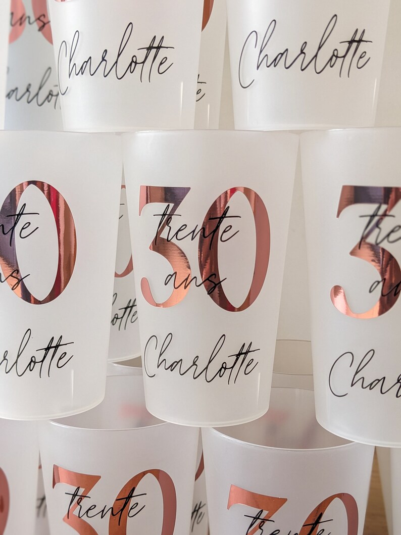 May include: A stack of white plastic cups with rose gold lettering. The cups are decorated with the name "Charlotte" and the number "30" with the text "trente ans" underneath.