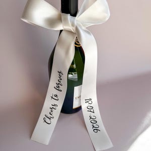 Könnte beinhalten: Eine Champagnerflasche, verziert mit einer großen weißen Satinschleife. Auf den Bändern der Schleife stehen die Worte "Cheers to forever" und das Datum "18.07.2026" in schwarzer Schrift. Die Flasche ist dunkelgrün mit schwarzem Verschluss.