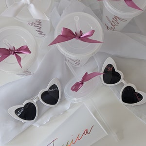 Op de afbeelding: Set van zes witte plastic bekers met roze strikken en rietjes. Elke beker heeft een naam erop geschreven:  "Camille", "Ashley", "future", "starlite", "evil", "carlisle".  Er is ook een witte beker met de naam "Jessica" erop geschreven in regenboogkleuren.