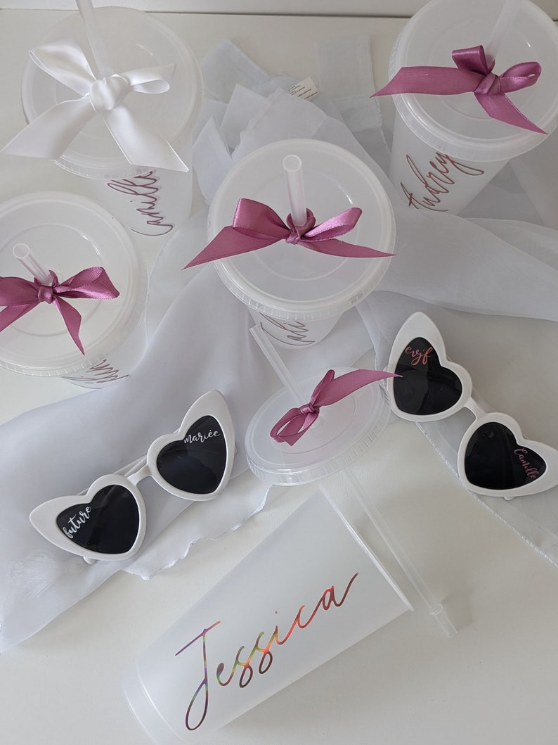 Op de afbeelding: Set van zes witte plastic bekers met roze strikken en rietjes. Elke beker heeft een naam erop geschreven:  "Camille", "Ashley", "future", "starlite", "evil", "carlisle".  Er is ook een witte beker met de naam "Jessica" erop geschreven in regenboogkleuren.