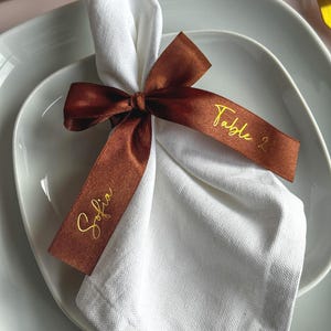 May include: A white linen napkin is folded and tied with a brown satin ribbon. The ribbon has gold lettering that reads "Sofia" and "Table 2". The napkin rests on a white plate.