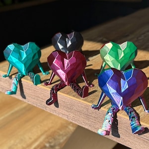 Könnte beinhalten: Fünf bunte, geometrische herzförmige Figuren mit Beinen, die auf einer Holzoberfläche sitzen. Die Herzen sind aus einem glänzenden, schillernden Material gefertigt und in einer Reihe angeordnet.