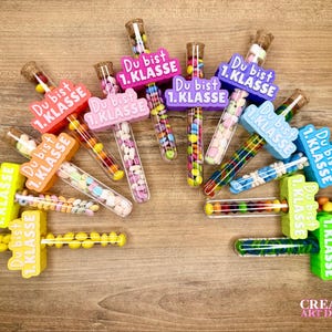 Könnte beinhalten: Eine Ansammlung von bunten, mit Süßigkeiten gefüllten Reagenzgläsern mit Korken. Jedes Reagenzglas hat einen farbenfrohen Kunststoffhalter mit dem Text "Du bist 1. Klasse". Die Süßigkeiten sind in verschiedenen Farben und Formen.