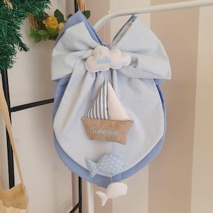 Può includere: Decorazione per la cameretta con un fiocco in tessuto a righe blu e bianche, una nuvola con la scritta "nato" e una barca a vela con il nome "Tommaso". Due ornamenti a forma di balena sono appesi sotto.