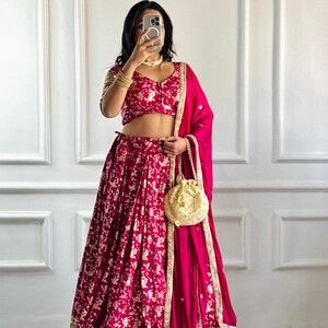 May include: A magenta lehenga choli set with a floral pattern. The set includes a crop top, a long skirt, and a matching dupatta. The skirt and top feature gold floral embroidery. A gold drawstring bag is also included.