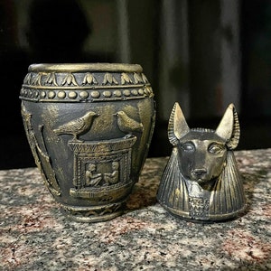 Op de afbeelding: Een zwarte en gouden keramische urn met een Egyptisch ontwerp. De urn heeft een afbeelding van twee vogels, een hiërogliefenpatroon en een scène met twee zittende figuren. De deksel is gevormd als een Anubis-hoofd.