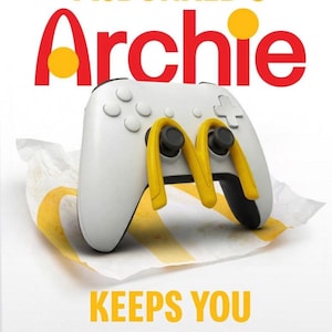 以下が含まれることがあります： 白いゲームコントローラー、ジョイスティックの代わりに黄色のマクドナルドのアーチが配置され、紙の包装紙の上に置かれています。「McDonald's Archie」の文字が上にあり、下に「KEEPS YOU IN THE GAME」と書かれています。
