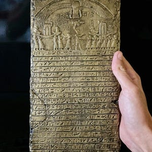 Könnte beinhalten: Eine goldfarbene Steintafel mit alten ägyptischen Hieroglyphen, die in die Oberfläche geschnitzt sind. Die Tafel zeigt eine geflügelte Sonnenscheibe und eine Prozession von Figuren.