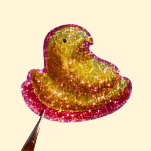 May include: A glittery yellow and pink chick-shaped embellishment. The chick has a rounded body and a small, curved head. The edges are outlined in a darker pink, and the body is covered in sparkling glitter. A pair of tweezers is holding the embellishment.