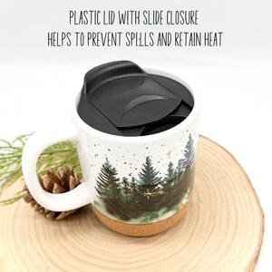 May include: White ceramic travel mug with a forest scene design. The mug has a black plastic lid with a slide closure. The text on the image reads: "PLASTIC LID WITH SLIDE CLOSURE HELPS TO PREVENT SPILLS AND RETAIN HEAT."