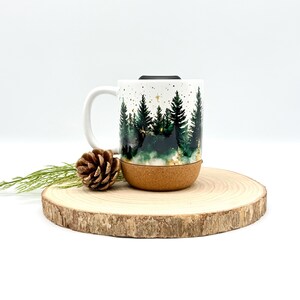 May include: White ceramic mug with a forest scene design. The mug features a handle and a cork base. The forest design includes green trees and gold stars. A pine cone and greenery are on a wooden slice next to the mug.