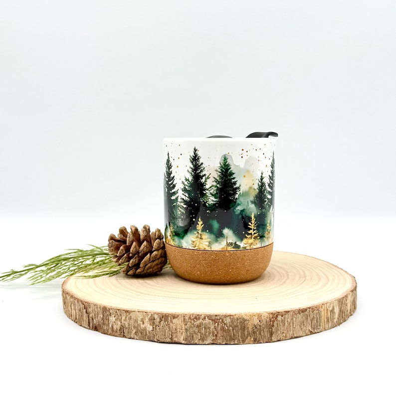 May include: White travel mug with a forest scene in shades of green and gold. The bottom of the mug is cork. A pine cone and greenery are next to the mug. The mug has a black lid.