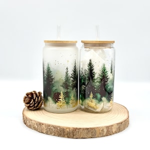 May include: Two glass tumblers with bamboo lids and clear straws. The tumblers feature a forest scene with green trees and gold accents. One is frosted, the other clear. They sit on a wooden slice with a pinecone.