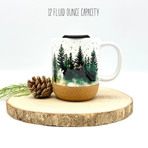 May include: White ceramic mug with a forest scene in shades of green and gold, with a cork base and black lid. The mug has a capacity of 12 fluid ounces. A pine cone and greenery are next to the mug on a wooden slice.