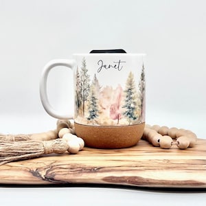 May include: White travel mug with a forest scene design and the name "Janet" in script. The bottom of the mug is wrapped in cork. The mug has a black lid and a white handle.