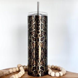 May include: A stainless steel tumbler with a gold and brown snake skin pattern. The tumbler has a lid and a straw.
