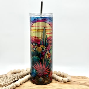 May include: A stainless steel tumbler with a colorful stained glass design of a desert sunset with cacti and flowers. The tumbler has a lid and a straw.