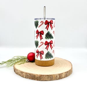 May include: A white tumbler with a Christmas-themed pattern featuring red bows, green Christmas trees, and candy canes. The tumbler has a stainless steel straw and a cork base. The tumbler is approximately 8 inches tall.