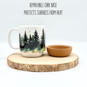 May include: A white ceramic mug with a forest scene design, a removable cork base, and a small cork coaster. The mug features a handle and is set on a wooden slice. Text on the image reads: REMOVABLE CORK BASE PROTECTS SURFACES FROM HEAT.