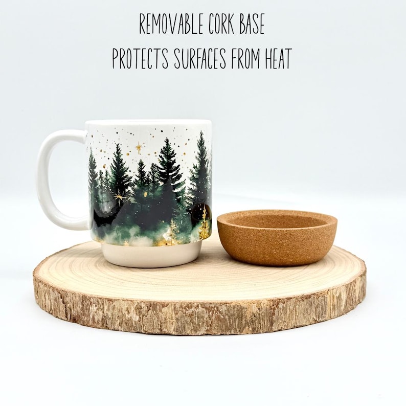 May include: A white ceramic mug with a forest scene design, a removable cork base, and a small cork coaster. The mug features a handle and is set on a wooden slice. Text on the image reads: REMOVABLE CORK BASE PROTECTS SURFACES FROM HEAT.