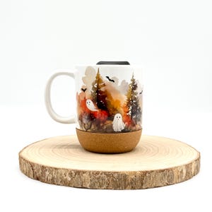 May include: White ceramic mug with a Halloween-themed design. The mug features a forest scene with ghosts, bats, and autumn colors. The bottom of the mug has a cork base. The mug is sitting on a wooden slice.