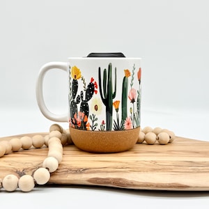 May include: White ceramic travel mug with a cork base and black lid. The mug features a colorful desert cactus and floral design. The handle is white. The mug is on a wooden cutting board with a strand of wooden beads.