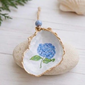 May include: A hand-painted oyster shell ornament featuring a blue hydrangea flower with green leaves. The shell has a gold-painted edge and a blue bead with a jute string for hanging. The ornament rests on a light-colored stone.