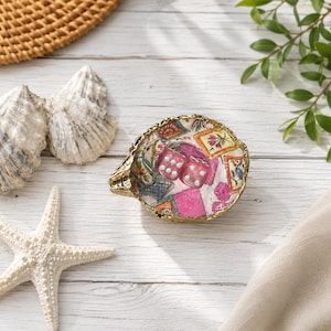 May include: A decorative oyster shell trinket dish with a gold-painted rim, featuring a colorful decoupage pattern of floral squares and pink textures, containing two translucent pink dice with white pips resting inside the shell bowl.