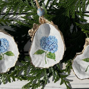 May include: Three oyster shell ornaments with a blue hydrangea design. The shells have a gold-painted edge and are strung with twine and a blue bead. The ornaments are displayed on a bed of green foliage.