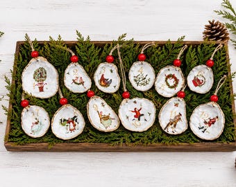 May include: A wooden tray holds twelve oyster shell Christmas ornaments. Each shell is hand-painted with a unique holiday scene and a number from 1 to 12. The ornaments are strung with twine and red beads, resting on a bed of greenery.