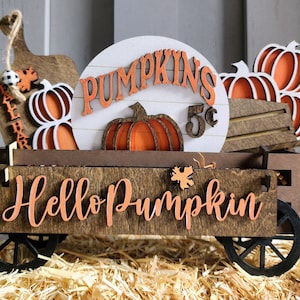 May include: Wooden fall-themed wagon with orange pumpkins and the words "Hello Pumpkin" and "Pumpkins 5¢". The wagon has black wheels and is set on a bed of hay. A decorative fall-themed item.