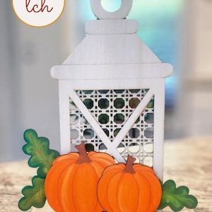 May include: White wooden lantern with a lattice design and two orange pumpkins on a wooden base. The lantern has a white handle with a circle on top.