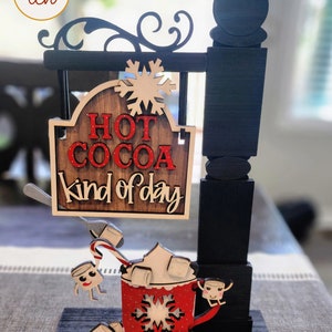 May include: Wooden sign with the words "HOT COCOA kind of day" in red and white. The sign features a red mug with whipped cream, marshmallows, and a candy cane. A snowflake and decorative scrollwork are also included.