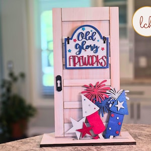 May include: Wooden patriotic door decoration with the words "Old Glory Fireworks." The door has a keyhole and is decorated with red, white, and blue fireworks and stars. The decoration is on a wooden base.