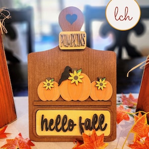 May include: Wooden fall-themed decoration featuring a brown cutting board shape. It has three orange pumpkins with yellow sunflowers, the word "PUMPKINS" above, and "hello fall" below. A blue heart is cut out at the top.