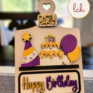 May include: Wooden birthday party sign with a purple and yellow party hat, a cake with candles, and balloons. The sign says "Happy Birthday" and "Party" in gold and purple lettering.
