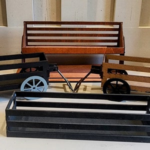 May include: Three wooden carts with wheels, two are black and one is brown. The carts are empty and have a slatted design. A wooden bench with a slatted back is behind the carts.