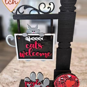 May include: A wooden cat-themed sign with a black frame and a gray cat cutout. The sign reads "cats welcome" in pink script, with a fishbone design. A paw print and a ball of yarn are also included.