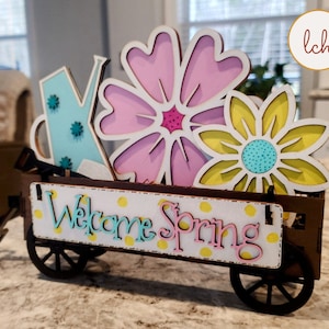 May include: A wooden wagon with wheels, decorated with a welcome spring sign, a blue watering can, a pink flower, and a yellow flower.