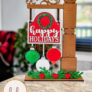 May include: Wooden holiday sign with a red background and the words "Happy Holidays" in white. The sign features a green wreath and three hanging ornaments in green, white, and red. A decorative base with green leaves and red berries completes the festive design.