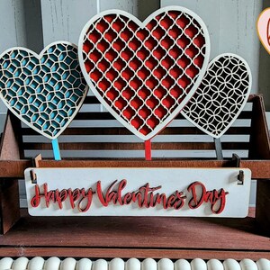May include: A wooden Valentine's Day decoration with three cutout hearts in red, blue, and black with a white background. The hearts are attached to a wooden stand with a white sign that reads "Happy Valentines Day" in red.