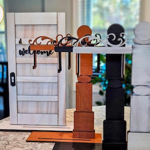 May include: Wooden welcome sign with a door design, featuring a decorative welcome sign and key hooks. The sign is accompanied by three decorative pillars in brown, black, and white. The sign is made of wood.