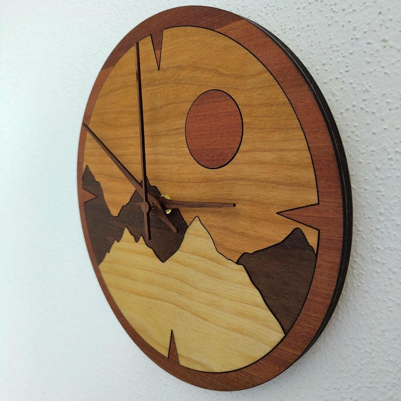 May include: A round wooden clock with a mountain and sun design. The clock face features a light brown background with a dark brown mountain range at the bottom and a red sun. The clock hands are dark brown.
