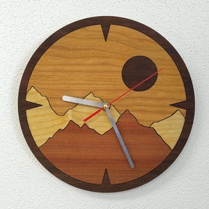 May include: A round wooden wall clock with a mountain landscape design. The clock face features a sun, mountains, and a red second hand. The clock is made of wood and has a dark brown frame. The clock hands are silver.