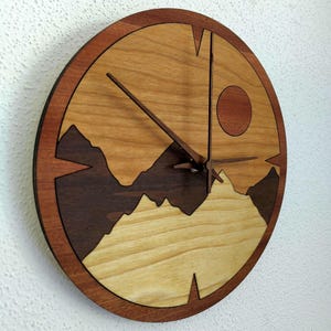 May include: A round wooden wall clock with a mountain and sun design. The clock face features a light wood background with a dark brown mountain range silhouette. The clock hands are dark brown, and the outer frame is a darker wood.
